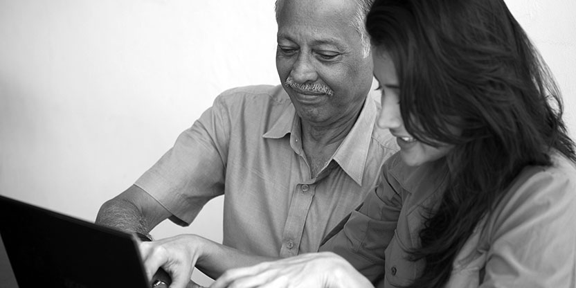 A young woman helping an elderly man to use the keyboard on a laptop.