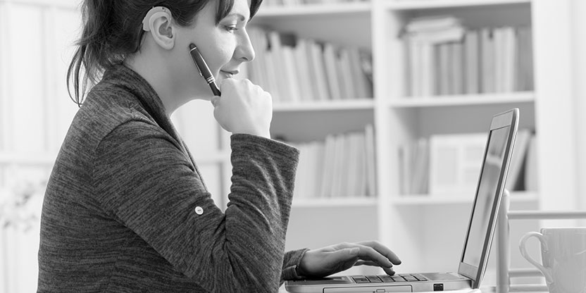 A woman in a home office wearing a hearing aid, uses her laptop.