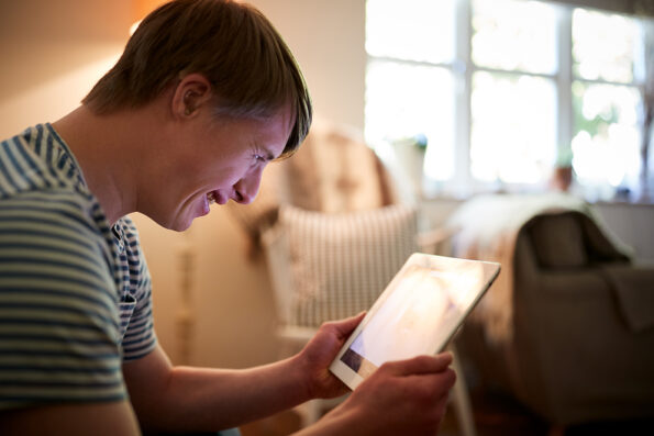 A young man with down syndrome and wearing a hearing aid smiles as he interacts with a tablet.